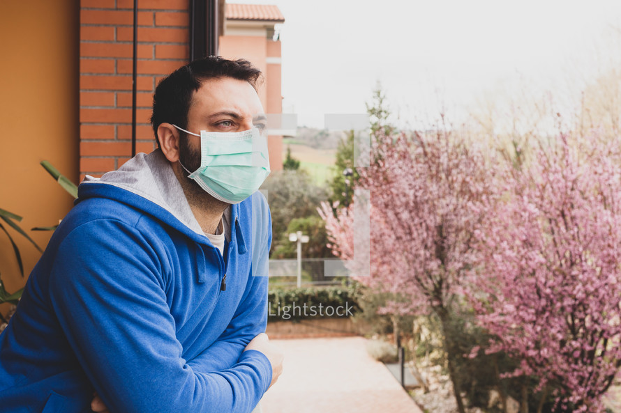 caucasian man with mask looking out on the terrace of the house during the quarantine due to the pandemic of covid19 coronavirus.