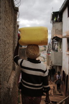 Person carrying water jug on their head through narrow alley