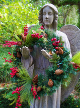 Angel statue holding a Christmas wreath  surrounded by green trees and forest accented by red berries, pine cones and a green wreath made out of pine tags and evergreen symbolizing the joy of the Christmas season in a tropical setting. 
