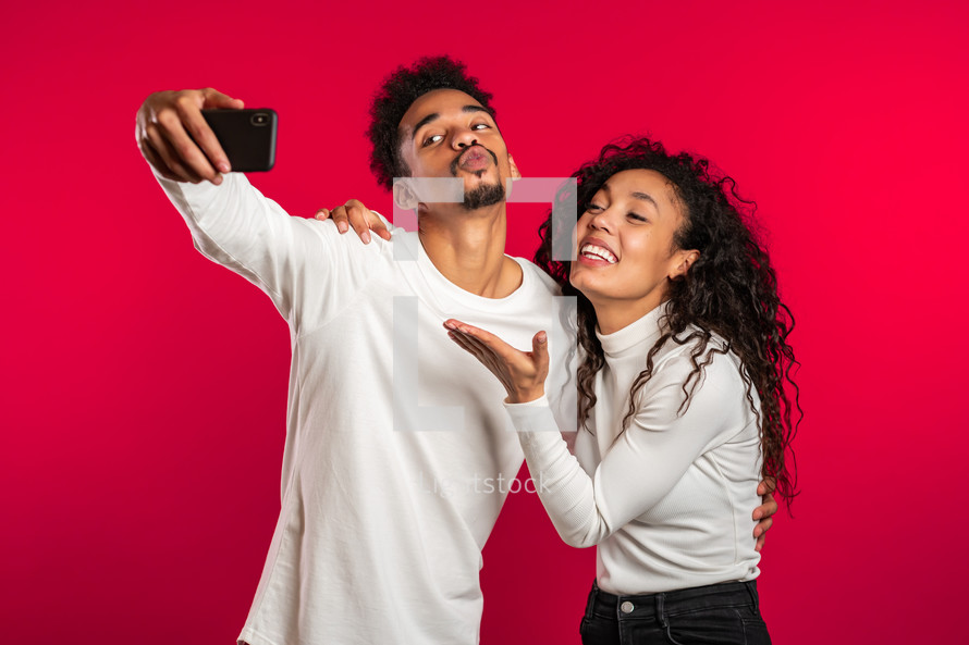 Young african american couple in white making selfie with smartphone on red studio background.Lovers match, love, holidays, happiness concept.