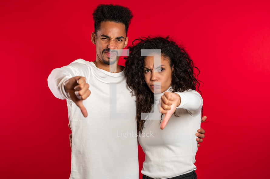 Young african couple standing on red studio background expressing discontent and showing thumb down gesture at camera. Portrait of man and woman with sign of dislike