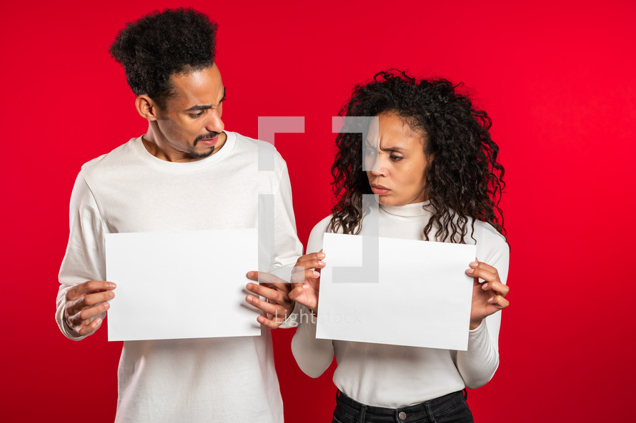 Portrait of young displeased african american man and woman couple holding white empty paper blanks on red studio background. Copy space. Match with separate sign.