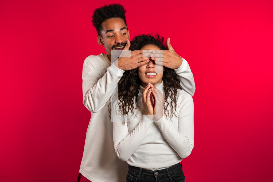 African man closes eyes of his beloved girlfriend before surprise her. Couple in white on red studio background. Love, holiday, happiness concept