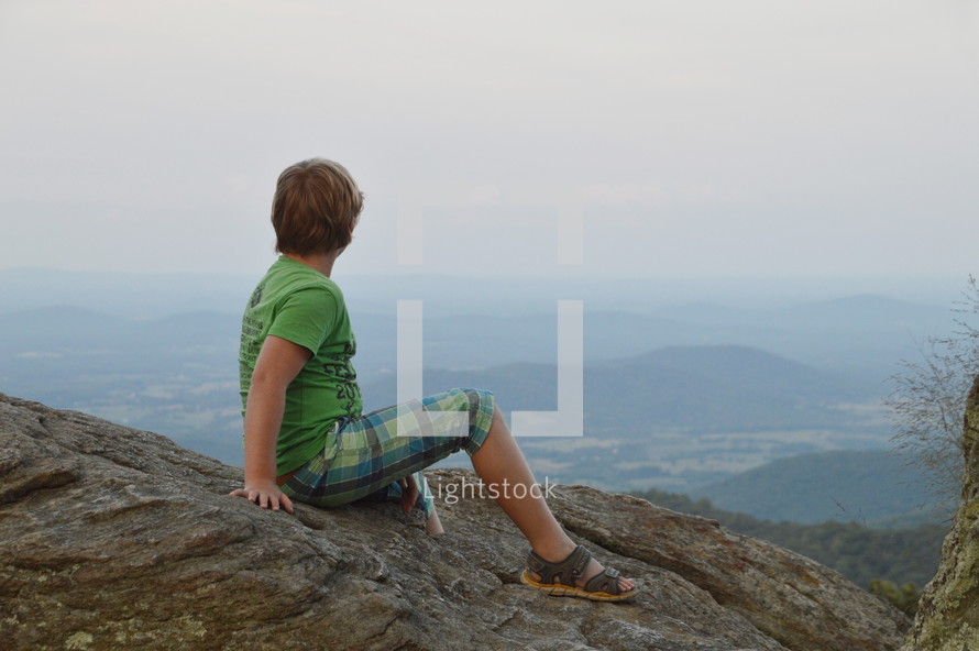 a boy sitting on a mountaintop taking in the view 