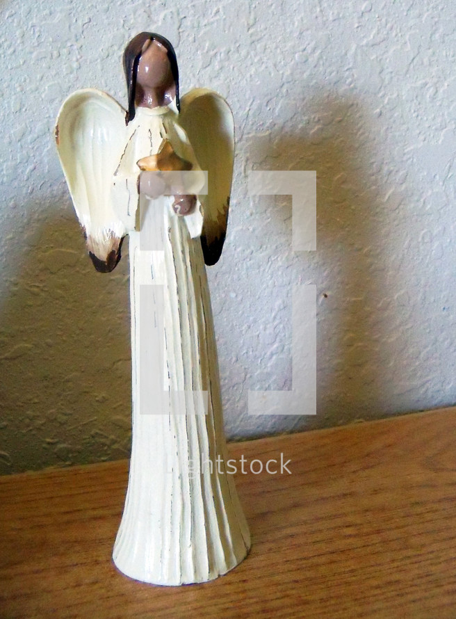 An Angelic Figurine sits on top of a wooden table against a stucco wall. 