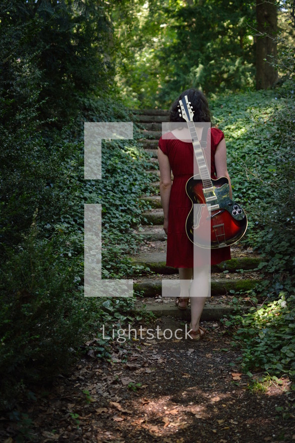 a woman in a red dress with an electric guitar walking through the dark woods