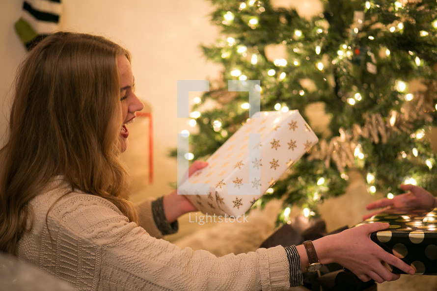 A laughing young woman holding gifts near a Christmas tree.