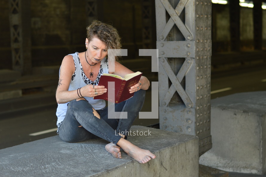 a punk woman sitting under a bridge reading a Bible 