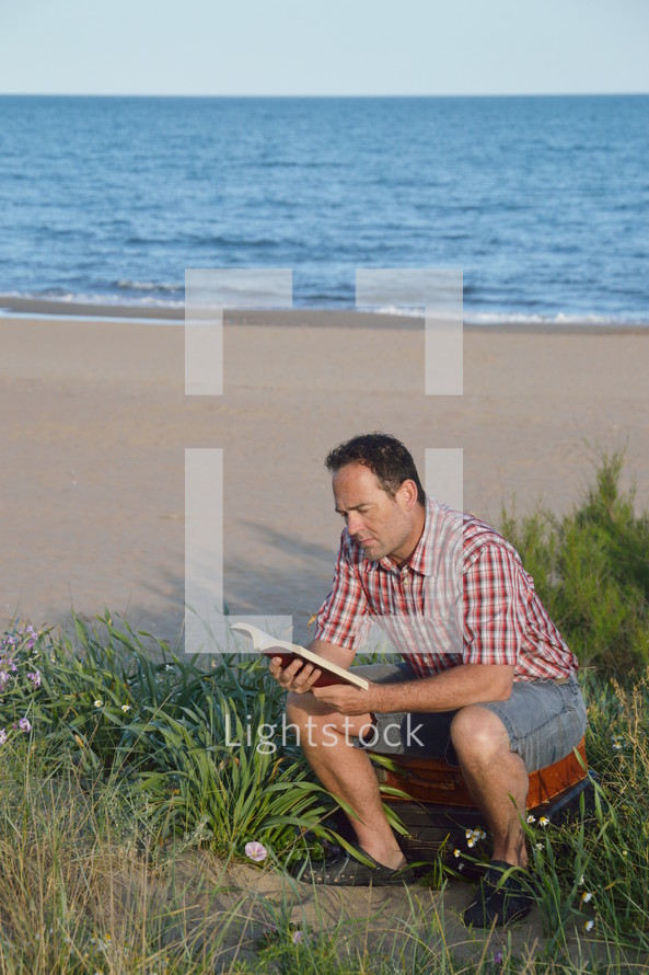 a man reading a Bible sitting on suitcase on a beach 