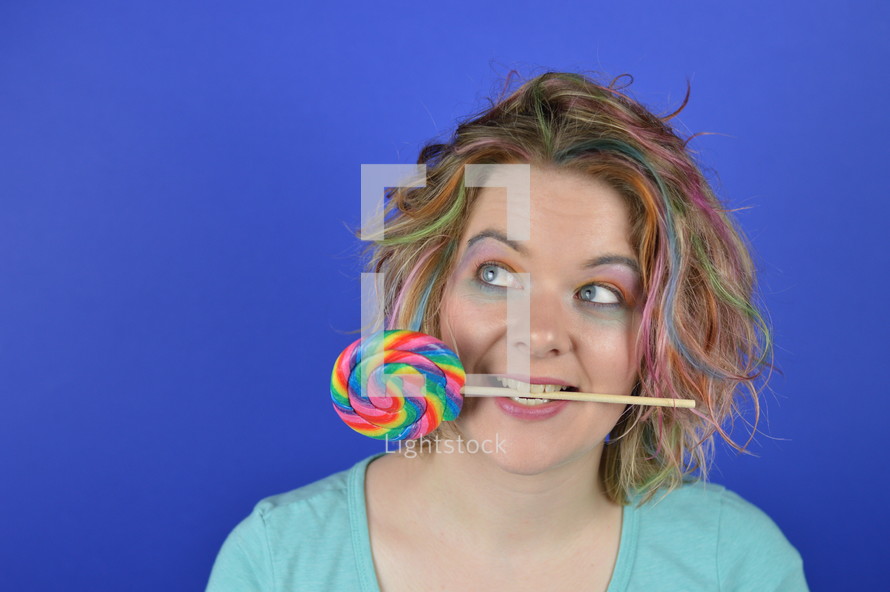 portrait of a young blond woman with colourful hair and a big lollipop between her teeth having fun and looking to the left corner above