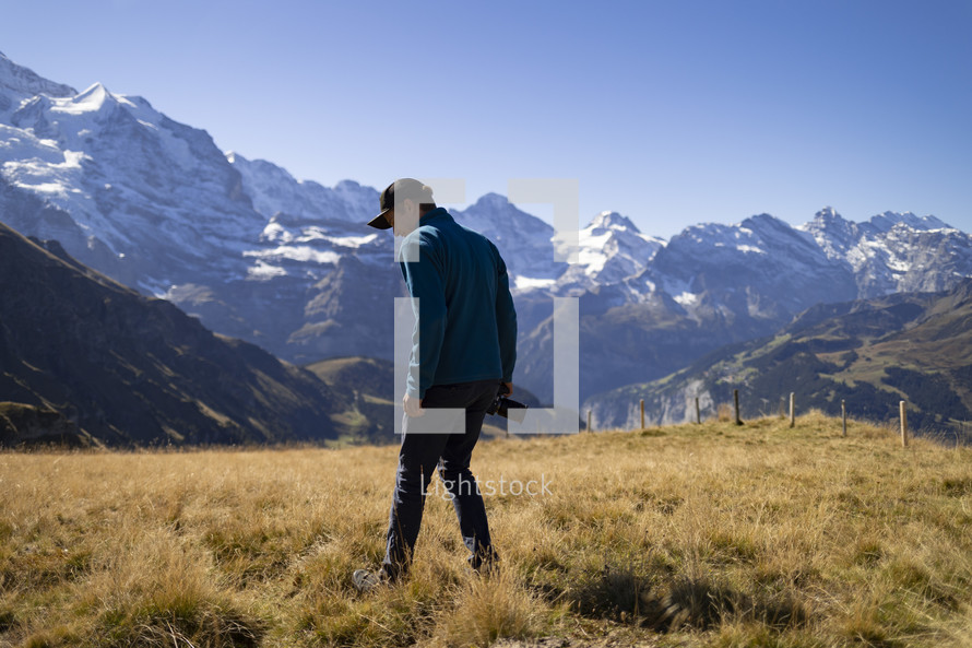 Man with camera in a grassy field in front of mountains with snow