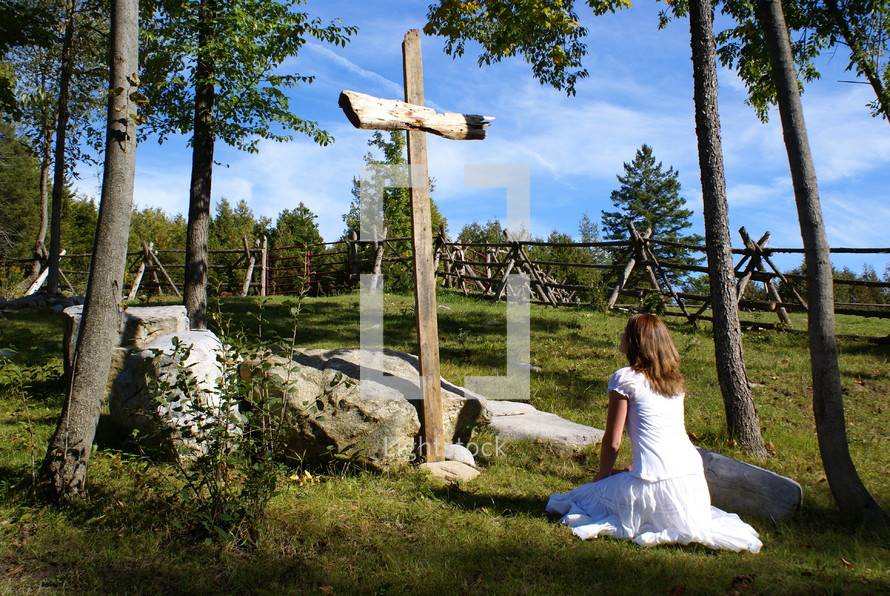 woman kneeling in prayer in front of a wood cross outdoors 