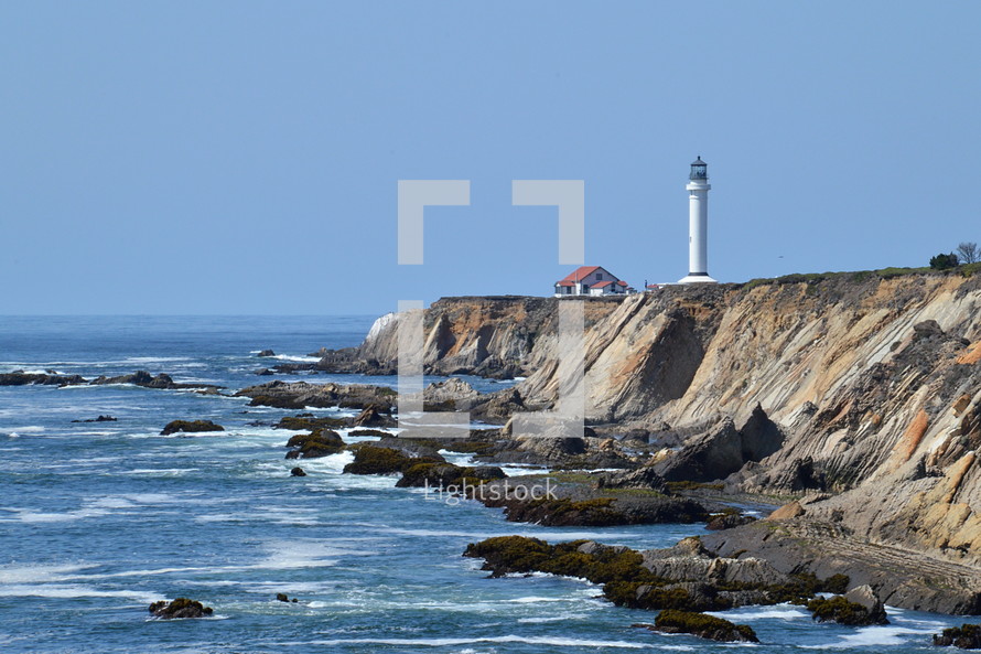 Lighthouse at the beach. 
lighthouse, shore, ocean, sea, light, white, shipping, navigation, boat, safe, secure, reliable, preservation, ship, ships, sky, blue, horizon, skyline, rocks, rock, stone, stones, cliff, cliffs, danger, dangers, dangerous, warning, caution, premonition, advance notice, advisory, caveat, admonition, warn, alert, admonish, save, safeguard, guard, rescue, protecting, protect, protection, coast, seaside, shoreline, seashore, seacoast, waterside, coastline, seafront, ragged, jagged, rugged, forbidding coastline, stony, craggy, bouldery, water, wave, waves, sand, house, tower, high