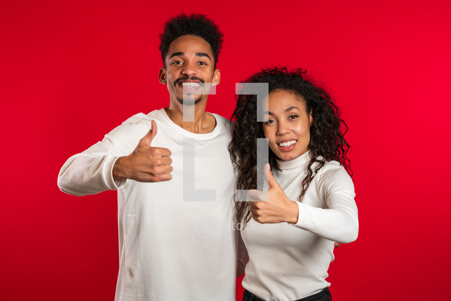 african american couple making thumbs up like sign over red background. Winner. Success. Positive girl and man smiles to camera. 