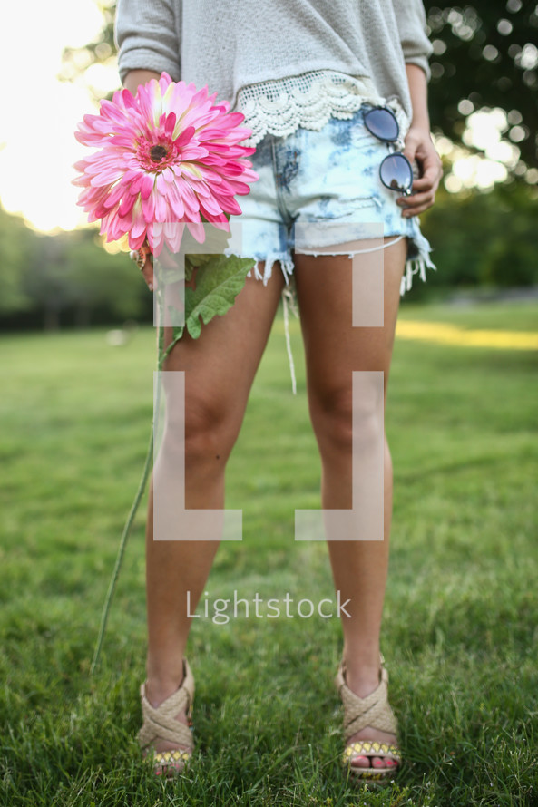 the legs of a young woman in shorts holding a flower and sunglasses 