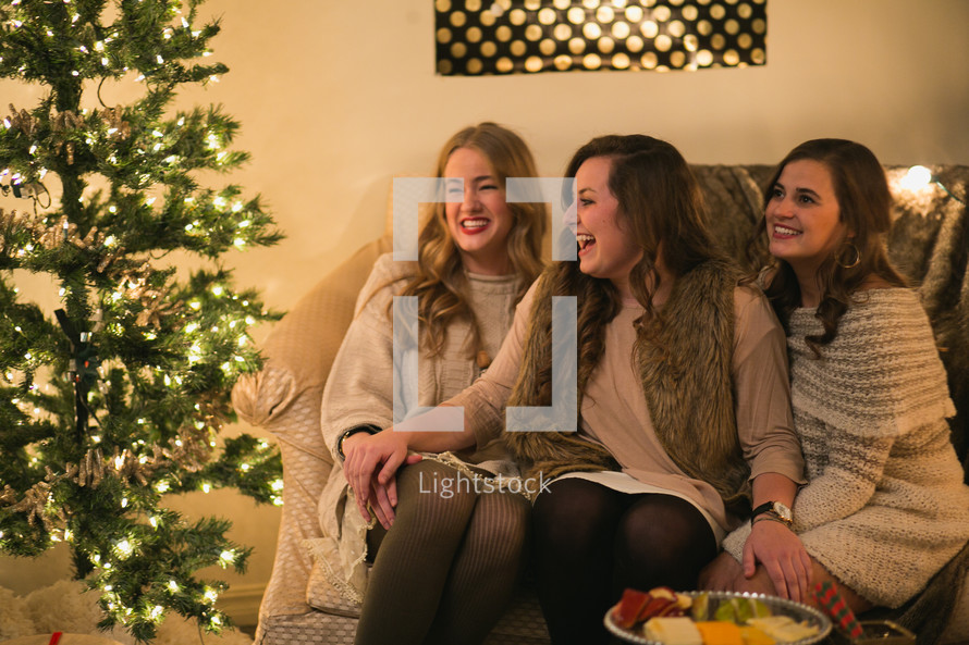 Three women sit together and laugh near a Christmas tree.