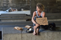 a woman sitting under a bridge with bottle of alcohol holding a sign 