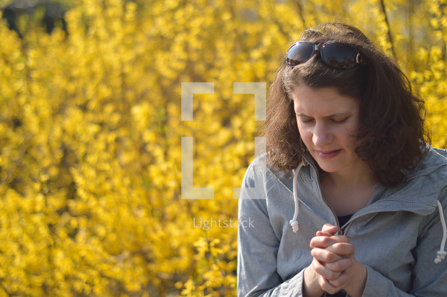 Young brunette woman praying with closed eyes while sitting outdoors in front of a bright yellow blooming forsythia bush. 