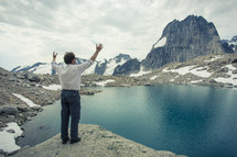 a man with raised hands standing at the edge of a lake surrounded by snow covered mountains 