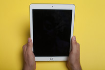 Hands holding an electronic tablet on a yellow background.