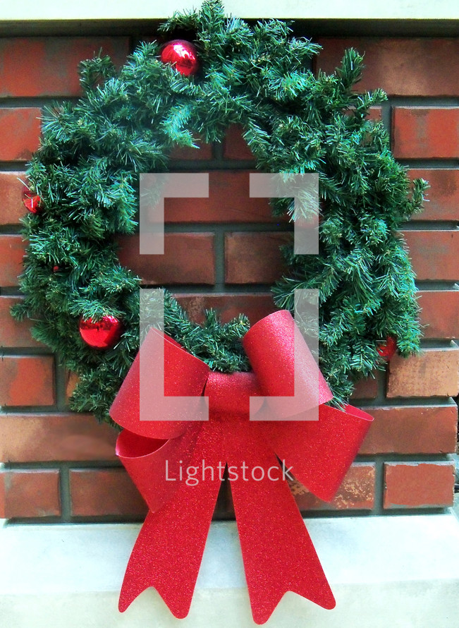A  traditional old fashioned green evergreen Christmas wreath with a red bow ribbon against a brick hearth background reminding viewers that the Christmas season is here. 