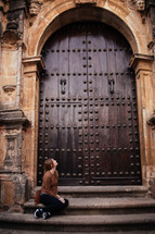 a woman sitting on steps looking up at large wooden doors at the entrance to a church 