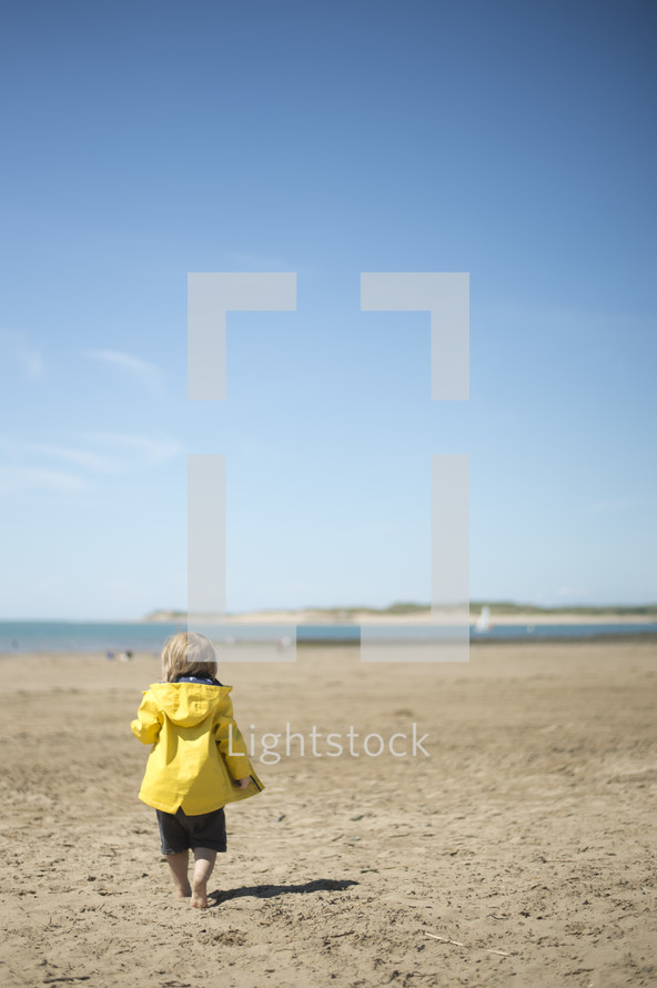 a toddler boy walking on a beach in a raincoat 