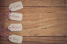 Christmas gift tags, reading "Love," "Hope," "Peace," and "Joy," on a wood grain background.