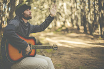 a man in a forest with a guitar and raised hand praising God 