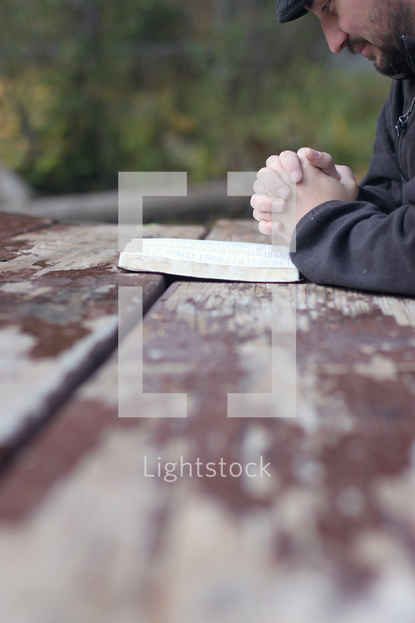 man praying near an open Bible on a picnic table 