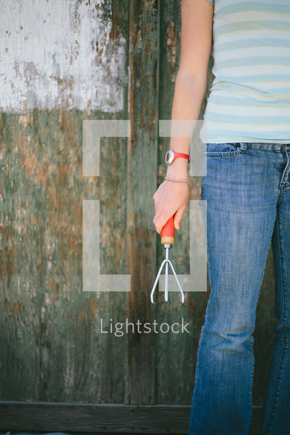 Teen holding a garden claw tool standing near a half painted fence.