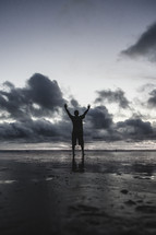a man standing on a beach on wet sand with hands raised at sunset 