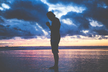 a man standing on a beach on wet sand with praying hands at sunset 