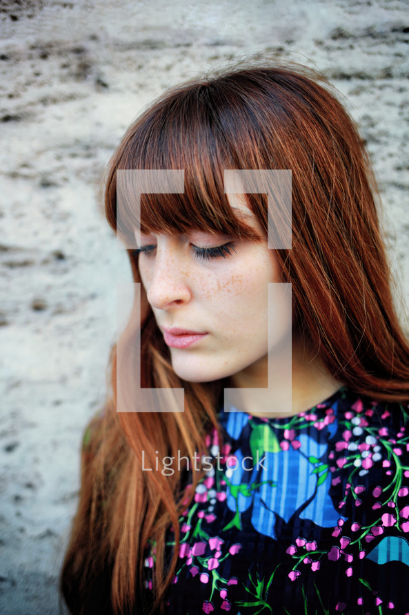 Young woman with long brown hair and freckles, wearing a floral print top, is looking down with a thoughtful expression