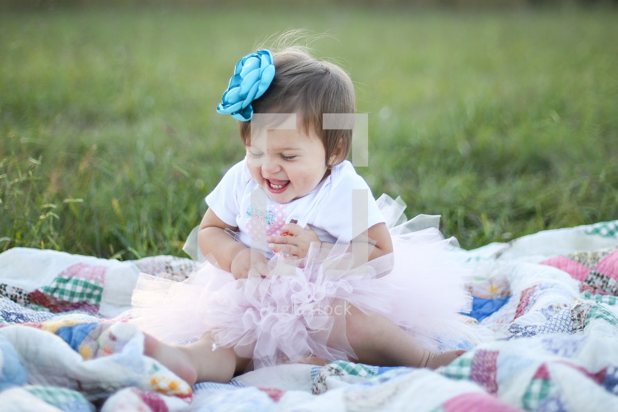 Smiling toddler girl in a tutu sitting on a quit in a field of grass.