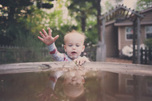 A baby boy plays in the water of a birdbath.