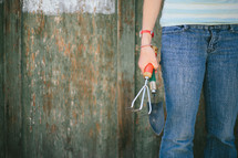 Teen holding a claw garden tool and spade shovel near a half painted fence.