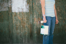 Young woman holding paint can and a paint brush, standing against an old wooden wall