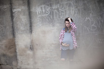 A pregnant teenager stands against a wall covered in graffiti.