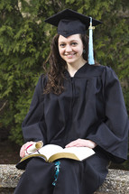 woman sitting outside in her cap and gown at graduation holding a Bible and a diploma