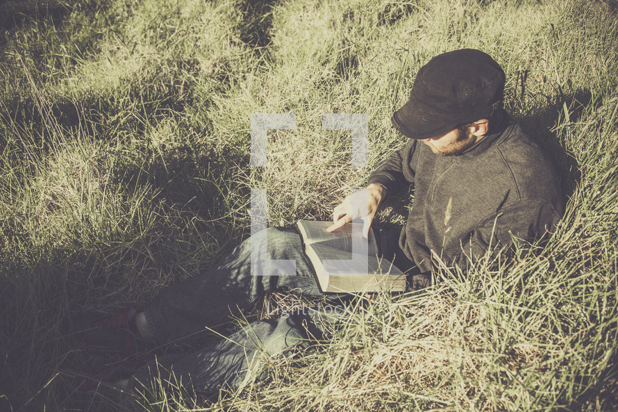  man sitting outdoors in tall grass reading a Bible  