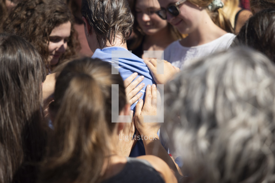 close up of hands and people surrounding and praying for a young woman who has been baptized by a lake 