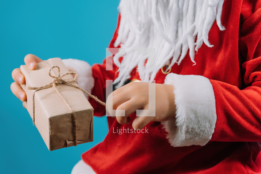 Hands of boy in Santa's costume pulling on string of wrapped gift box. Child wonders what is inside. Happy kid with present. Blue Christmas background. High quality photo