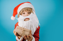 Excited boy in Santa costume with beard holding gift box with bow. Child, Happy kid with present. Blue Christmas background. Copy space, banner. High quality photo