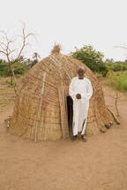 African man standing in front of a grass hut in a rural setting