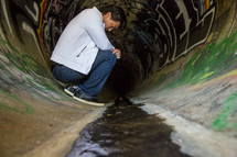 Man kneeling in a sewer drain pipe with graffiti.