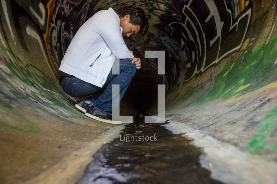 Man kneeling in a sewer drain pipe with graffiti.
