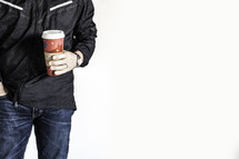 Man in jeans and black jacket with one hand in pocklet, holding red disposable coffee cup with the other.