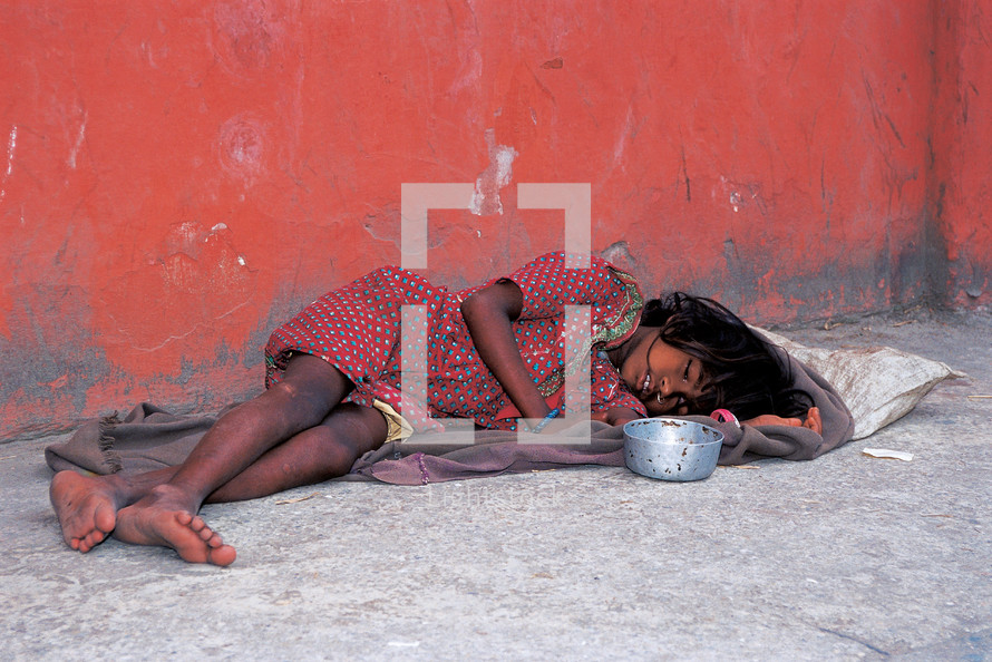 a homeless child sleeping on concrete with begging bowl