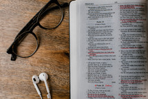 reading glasses, earbuds, and open Bible on wood table 